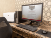 Computer workstation with a desktop PC, monitor, keyboard, mouse, and printer on a granite desk. The monitor displays a business center webpage. Black mesh chair in front.
