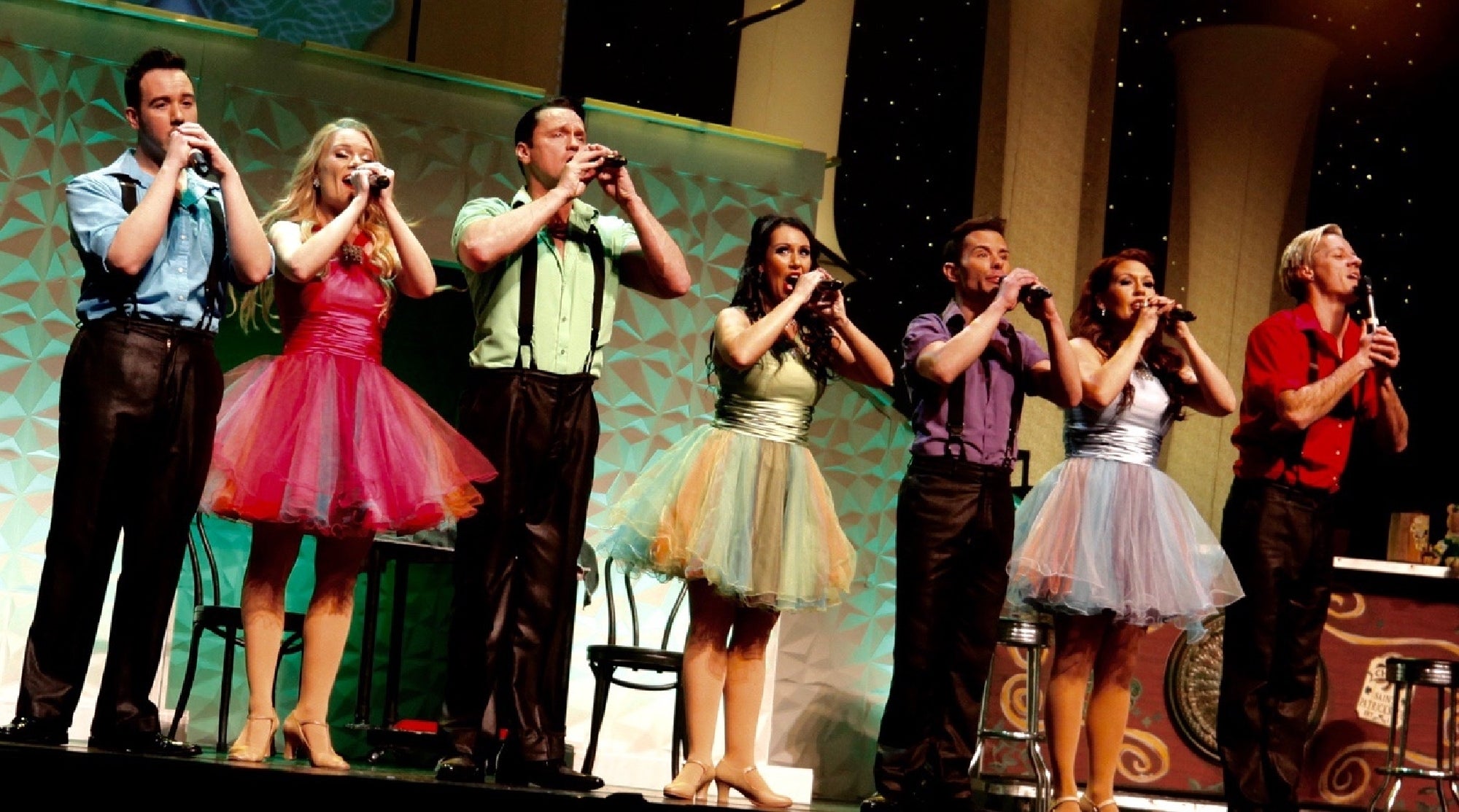 A group of performers on stage, each holding a microphone and wearing colorful outfits, singing in unison.