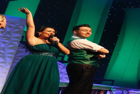 People on stage, one holding a microphone, both wearing green outfits, with a geometric backdrop and starry lights.