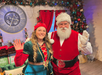 A person dressed as an elf and another as Santa Claus wave at the camera in a festive setting with Christmas decorations and a large clock on the wall.