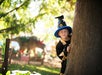 A child dressed in a wizard costume peeks out from behind a tree in a sunlit outdoor setting.