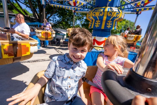 Two children smiling and enjoying a ride at an amusement park on a sunny day. Other people are visible in the background.