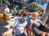 A young girl and boy smiling as they ride a carousel together, with other park guests enjoying the ride in the background on a sunny day.