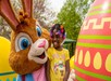 Child smiling next to a person in a bunny costume, standing by a large decorative Easter egg, with trees in the background.