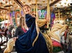 A child in a blue dress smiles while riding a carousel horse, with an adult holding her from behind. Bright lights and decorative details are visible in the background.