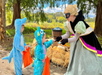Two children in blue animal costumes receive candy from a masked woman in a princess dress at an outdoor Halloween event with hay bales and autumn decorations.