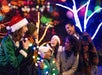 Six people in winter clothes and Santa hats stand outdoors at night, smiling and laughing together, surrounded by colorful holiday lights and illuminated trees.