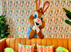 A person in a brown rabbit costume stands behind an orange couch with carrot-themed cushions against a carrot-patterned wallpaper.