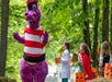 A person in a purple dinosaur costume interacts with three children in costumes holding pumpkin-shaped buckets in a wooded outdoor setting.