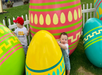 Two young children play around large, colorful decorative Easter eggs in a grassy area. A white picket fence is in the background.