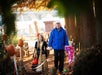 An older man walks outdoors holding hands with two young girls in costumes; one is dressed as a princess, the other in pink, with carved pumpkins along the path.
