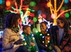 A family of four sits outdoors at night, smiling and talking, with colorful holiday lights illuminating trees in the background.