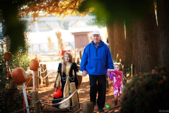 A girl in a costume holding a pumpkin bucket, walking hand-in-hand with an older man in a blue jacket, accompanied by a younger girl in a pink outfit, amidst a festive autumn setting decorated with pumpkins.