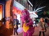 A person in a purple dragon costume poses with smiling children outside a brightly lit building at night, while another person takes their photo.
