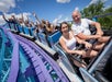 A group of people on a colorful roller coaster ride, smiling and raising their hands in excitement, under a bright blue sky with scattered clouds.
