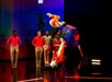 Performers in red and white costumes execute an acrobatic flip on stage, with several others standing in the background against a colorful set.