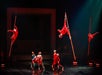 Performers in red costumes execute aerial and acrobatic acts on stage, with some suspended by ropes and others standing or crouching below, under dramatic lighting.