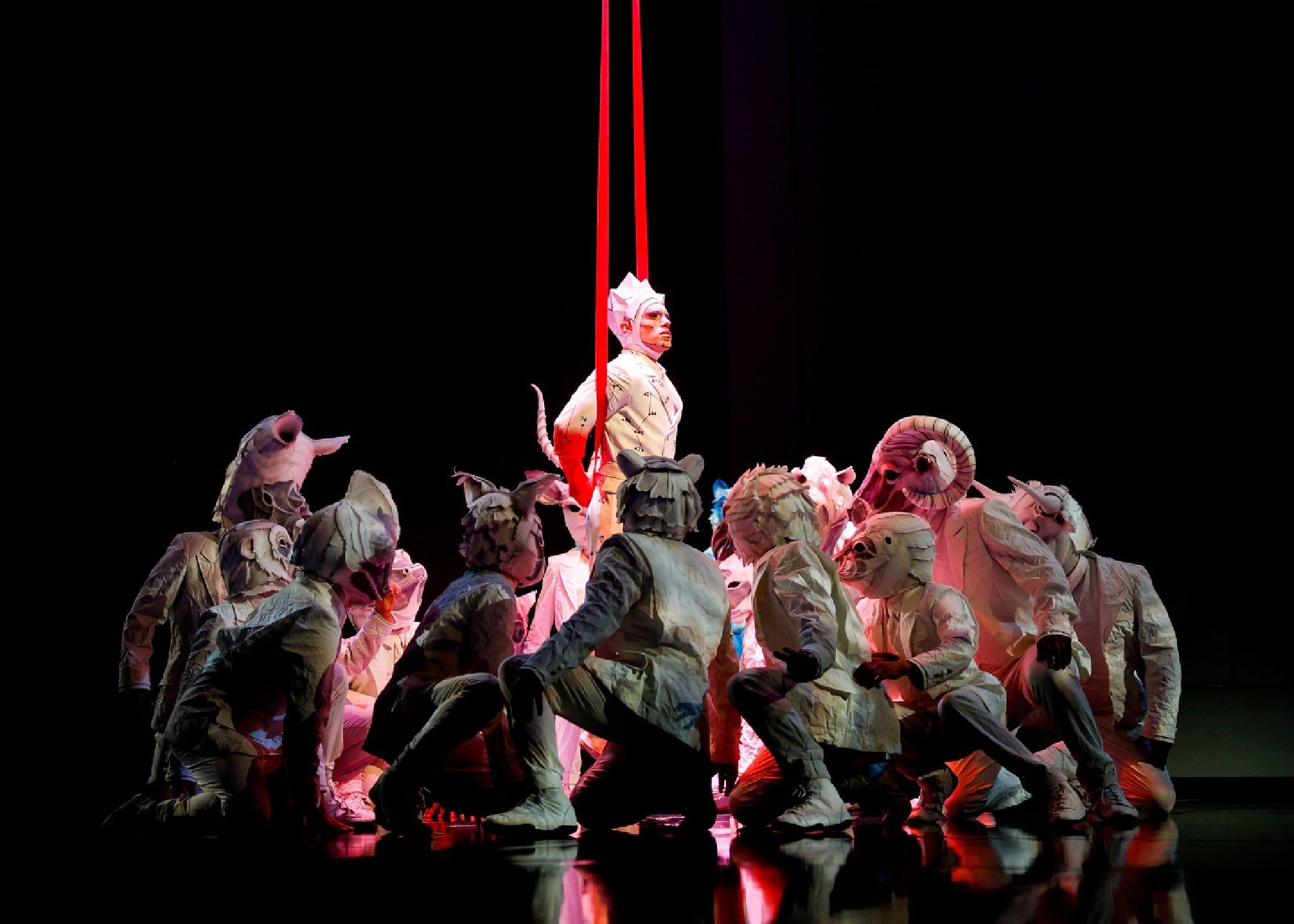 A group of performers in white animal costumes crouch around a central figure in white, who stands upright under two red vertical lights on a dark stage.