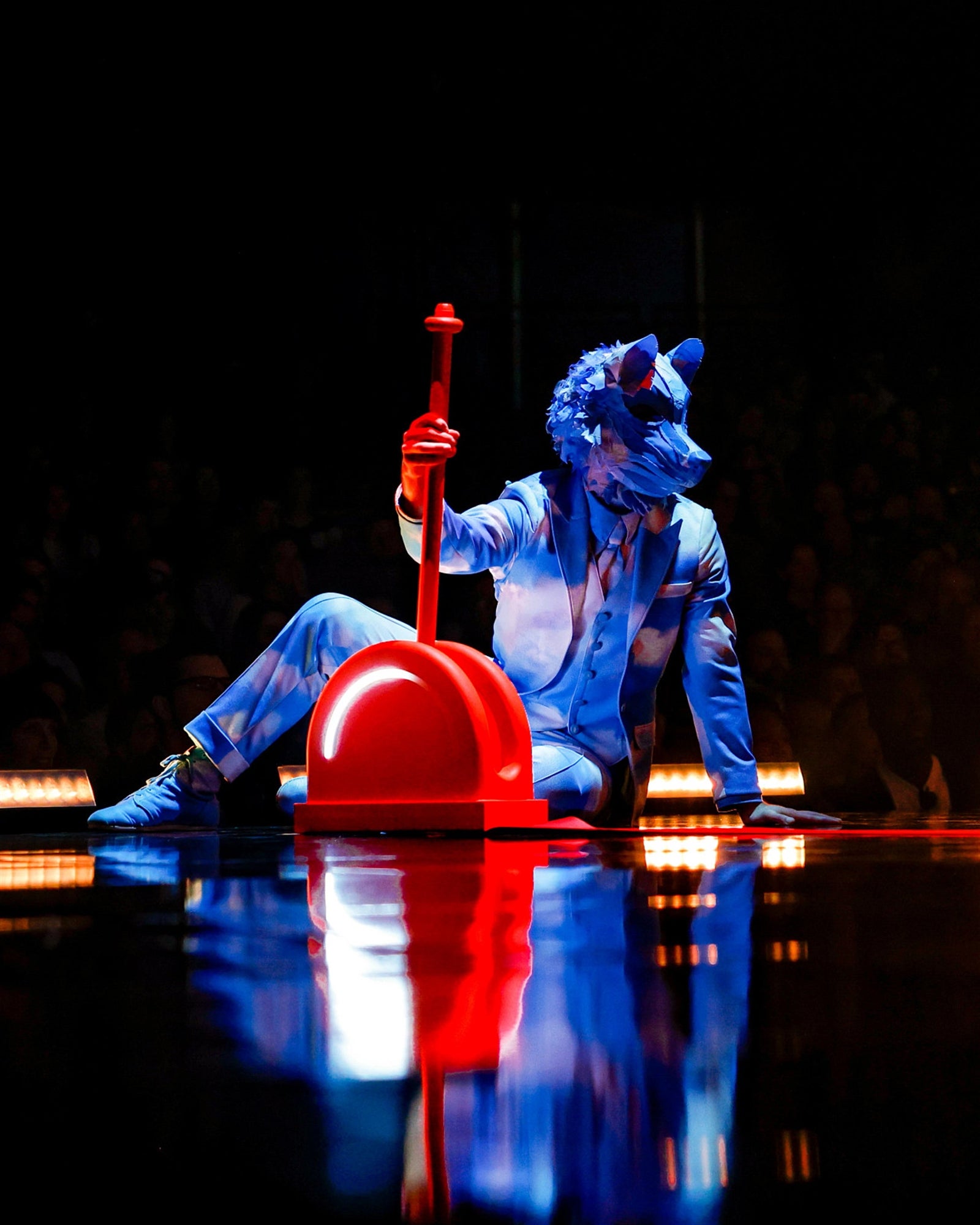 A person in a blue animal costume and suit sits on a glossy stage, holding a large red prop shaped like a shovel or lever, with dim lighting and a visible audience in the background.