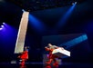 Two performers in red outfits balance and hold large white boxes on a stage with dramatic blue and purple lighting.