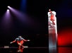 Two performers in red costumes are on stage; one stands with arms open at the base, while the other climbs a tall white stack, reaching upward under dramatic lighting.