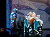Performers dressed in animal costumes, including a wolf and several with fish heads, act on a brightly lit stage with a blue and white backdrop.
