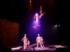 Acrobats in white costumes are mid-air above a seesaw-like apparatus on stage, performing a coordinated stunt under dramatic lighting.
