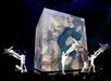 Performers in white costumes are suspended on wires around a large illuminated cube displaying an underwater scene, with an audience watching in the foreground.