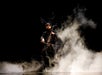 A cellist wearing a dark suit and antler headpiece plays on stage surrounded by dramatic smoke against a black background.