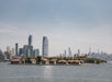 An island with historic buildings sits in the foreground, with the New York City skyline and tall modern skyscrapers in the background across the water.