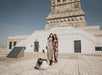 A person lies on the ground taking a photo of two women standing in front of a large stone monument under a clear sky.