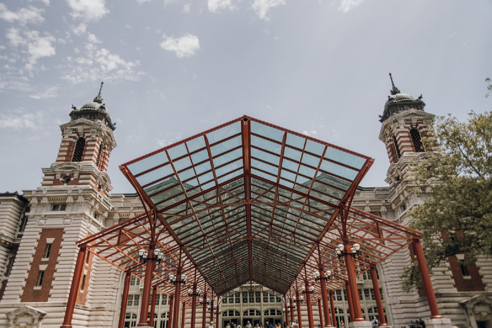 A large red metal and glass canopy extends from the entrance of a historic building with two clock towers and ornate architectural details under a partly cloudy sky.