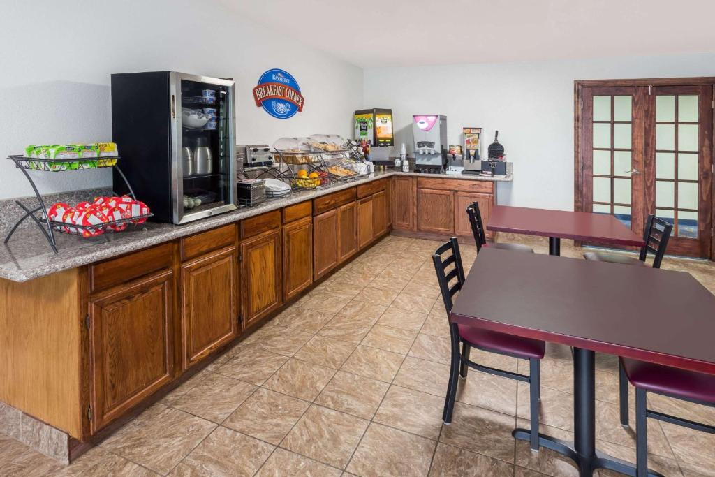 Hotel breakfast area with wooden cabinets, tables, and chairs. Features a coffee machine, cereal dispensers, juice machine, and various food items displayed on the counter.