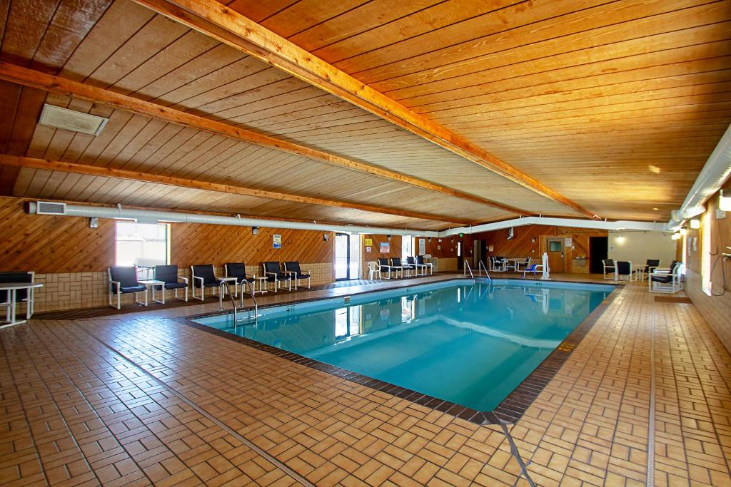 Indoor swimming pool with wooden walls and ceiling. Surrounding the pool are tables and chairs. Bright lights illuminate the area through windows and ceiling lights.