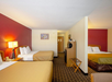 Hotel room with two double beds, red accent wall, wooden furniture, and a TV. A lamp and wall art are visible. A luggage stand and doorway are in the background.