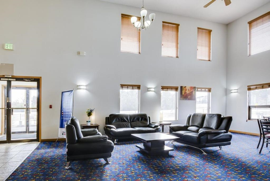 Spacious lobby with black leather sofas, a modern coffee table, and a colorful carpet. Large windows with blinds illuminate the area, and a chandelier hangs from the high ceiling.