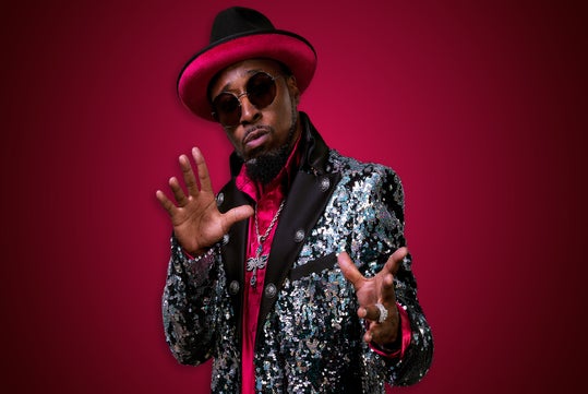 A man in a sequined jacket, red hat, sunglasses, and jewelry poses with his hands up against a red background.