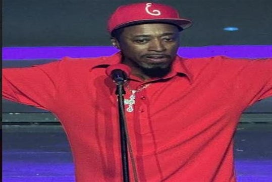 Person in a red shirt and cap speaking into a microphone on stage with arms outstretched.