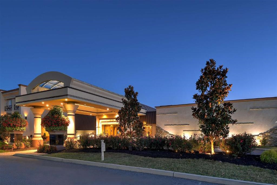 Exterior view of a well-lit building with columns, surrounded by landscaped gardens and trees, during twilight.