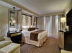 Luxurious hotel room with a large bed, brown and beige decor, two nightstands, armchair, and a TV. Floor-to-ceiling curtains cover wide windows.