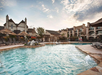 Outdoor pool area with lounge chairs and umbrellas in front of a two-story building under a partly cloudy sky.