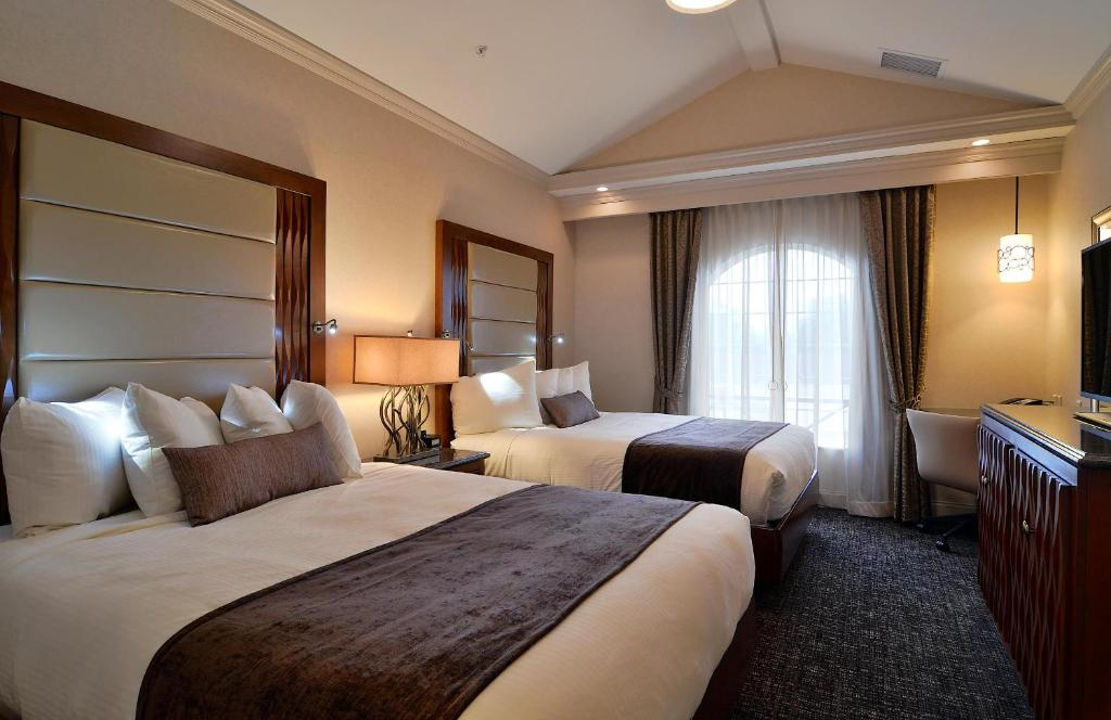 Hotel room with two double beds, a desk with a chair, a lamp, and a flat-screen TV. Large arched window with curtains illuminates the room.
