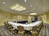 Conference room with U-shaped table setup, beige walls, and decorative ceiling lights. Chairs surround the table, which is set with water glasses and notepads. A TV is in front of draped curtains.