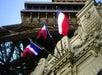Three French flags are displayed on a stone structure with architectural details, with part of the Eiffel Tower visible in the background.