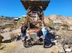Two people stand by an old rusty car in a desert setting with wooden structures and rocky hills in the background. A sign overhead reads "Hamm's.