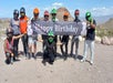 A group of nine people wearing helmets and casual clothes hold a "Happy Birthday" banner outdoors in a rocky, desert landscape.