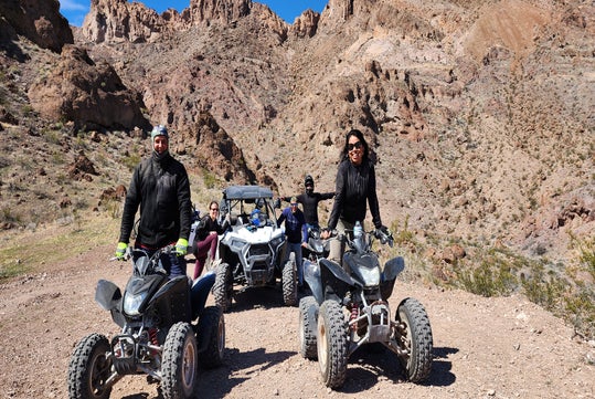 Four people riding two ATVs and a UTV on a rocky, mountainous terrain under a clear blue sky.