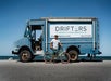 A person stands next to a vintage blue delivery truck with "Drifters Beach & Bike Rentals" signage, holding a green bicycle under a clear blue sky.