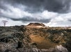 A barren volcanic landscape with dark clouds, rocky terrain, and steam rising from the ground. A lone, bare tree stands to the left.