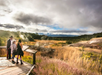 Three people stand on a wooden platform observing a geothermal landscape with steam rising from the ground, surrounded by grass and a cloudy sky.